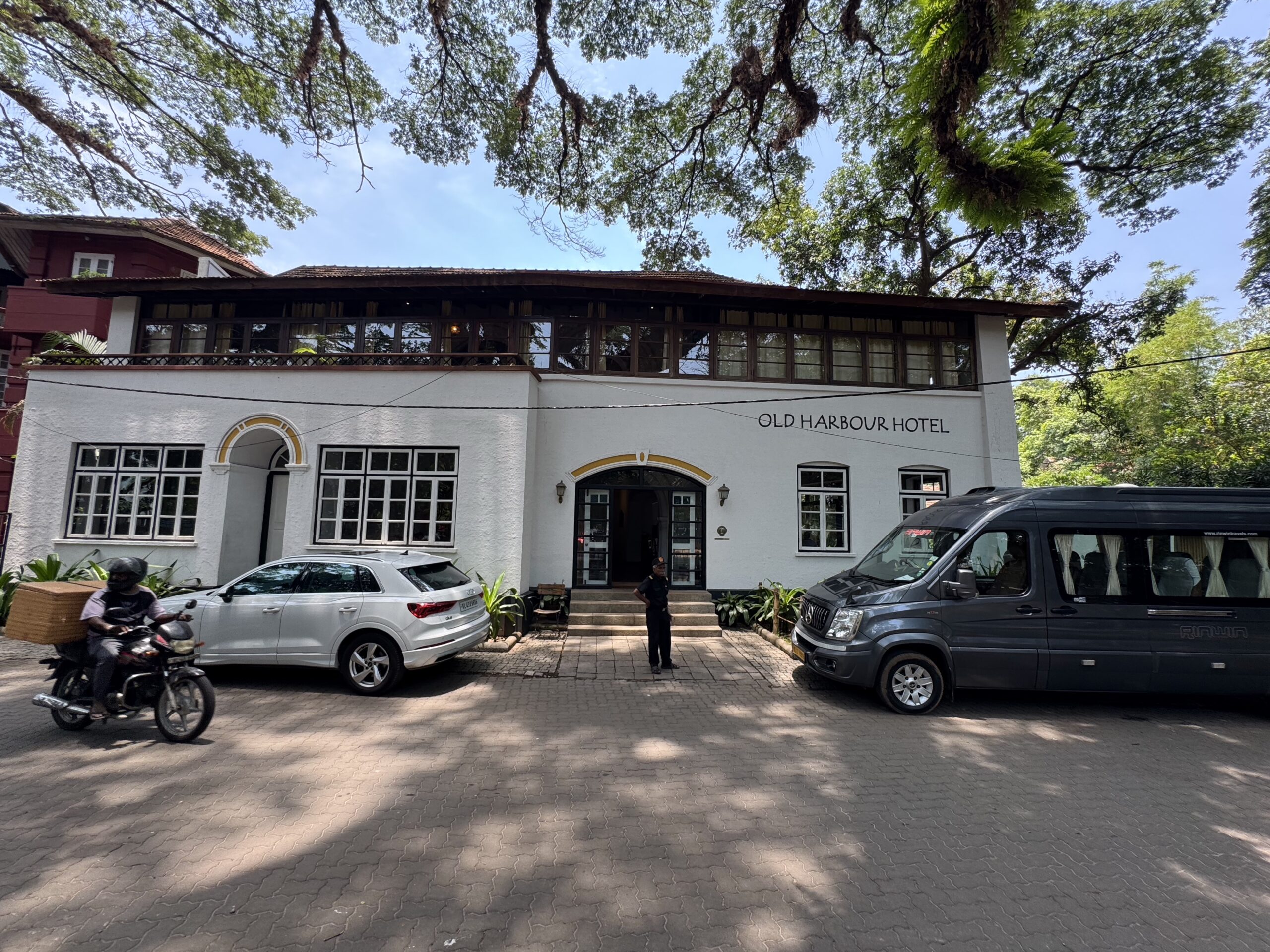 Front view of Old Harbour Hotel: white facade, arched entry, and parked cars in front under leafy trees.