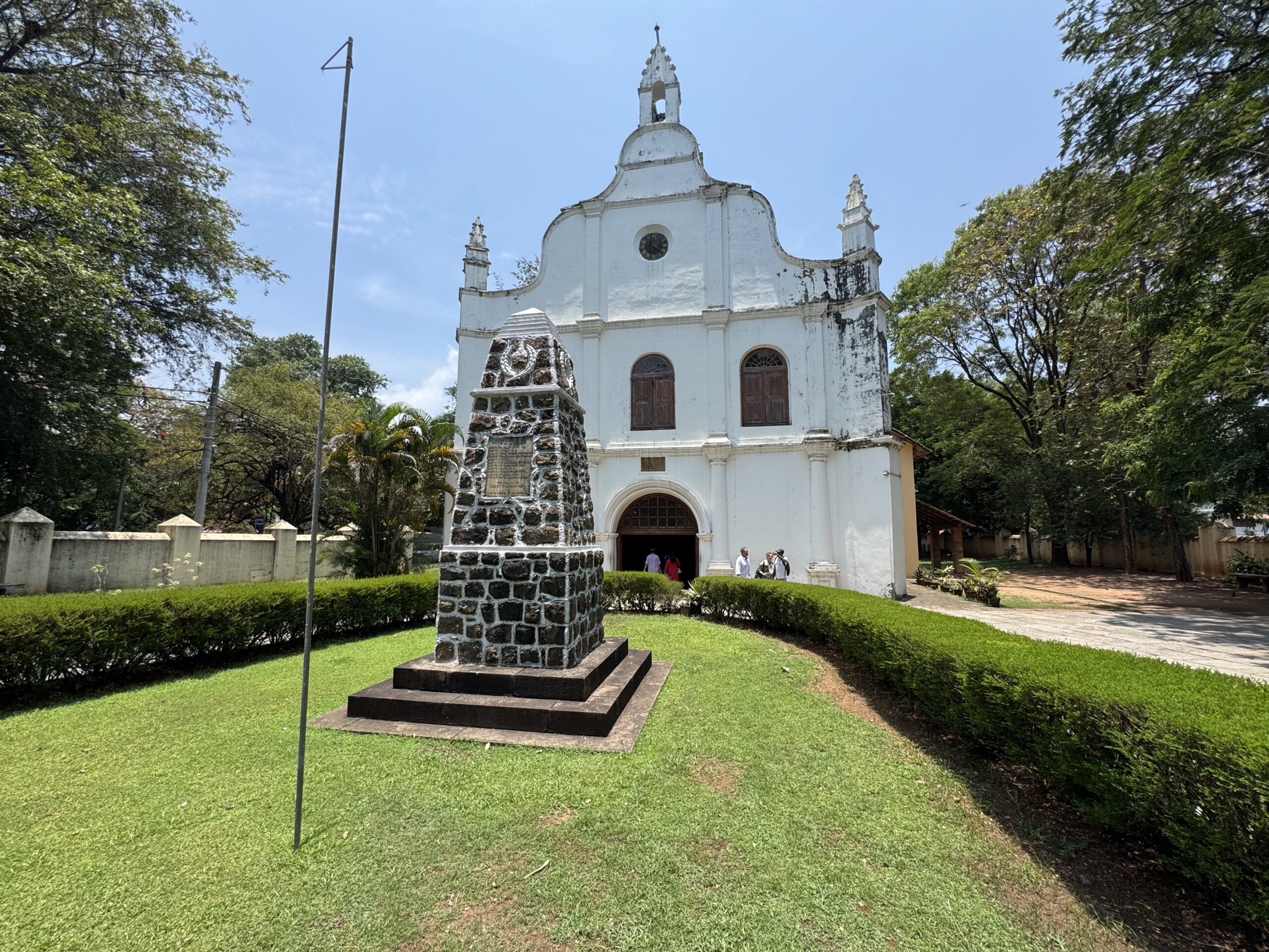 Front view of a white church with curved facade and small bell tower, set in a green park with a stone pedestal monument in the foreground.