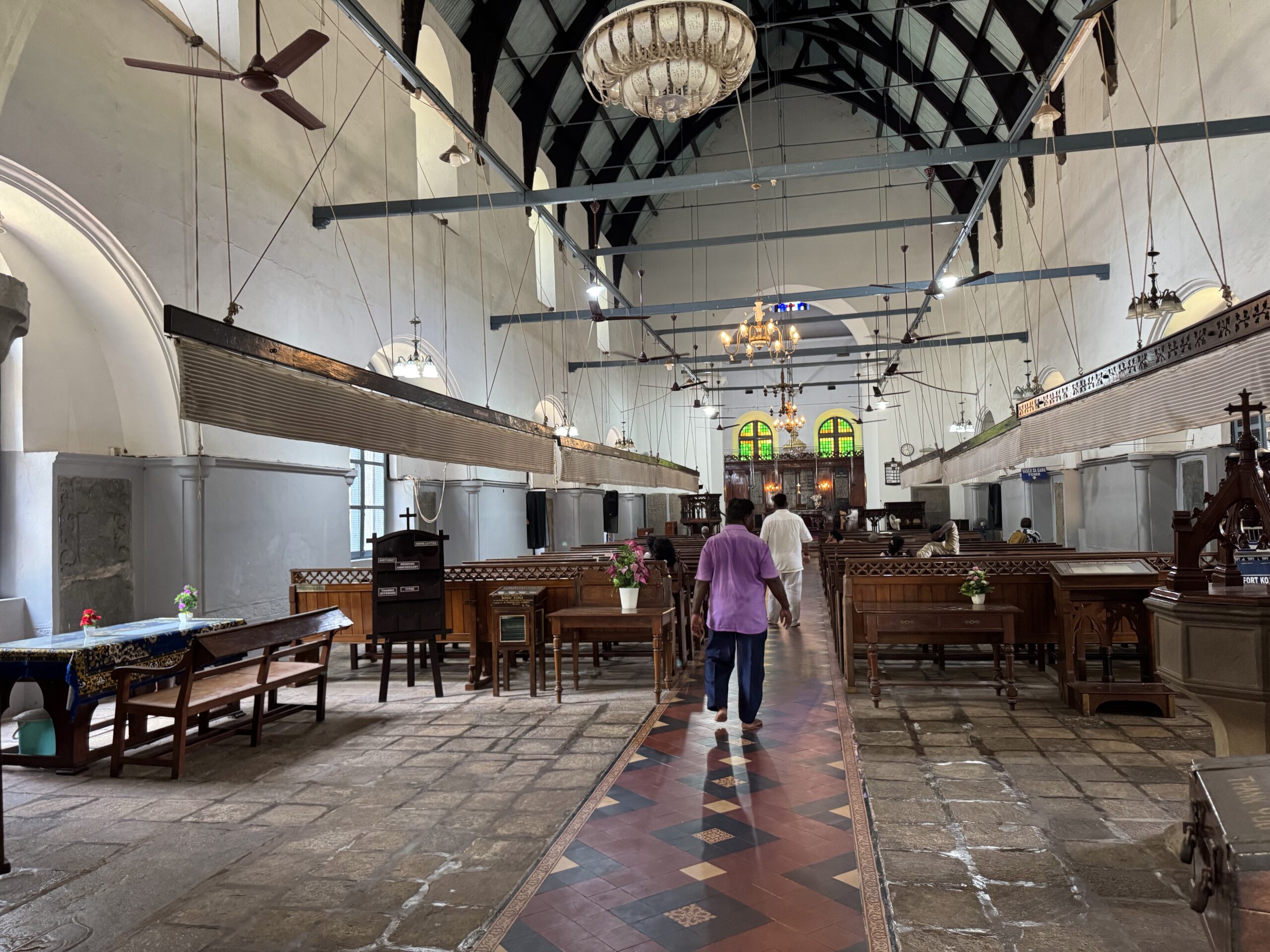 Interior of a church hall with wooden pews, hanging fans, chandeliers, and a painted tile floor; stained glass windows at the far end.