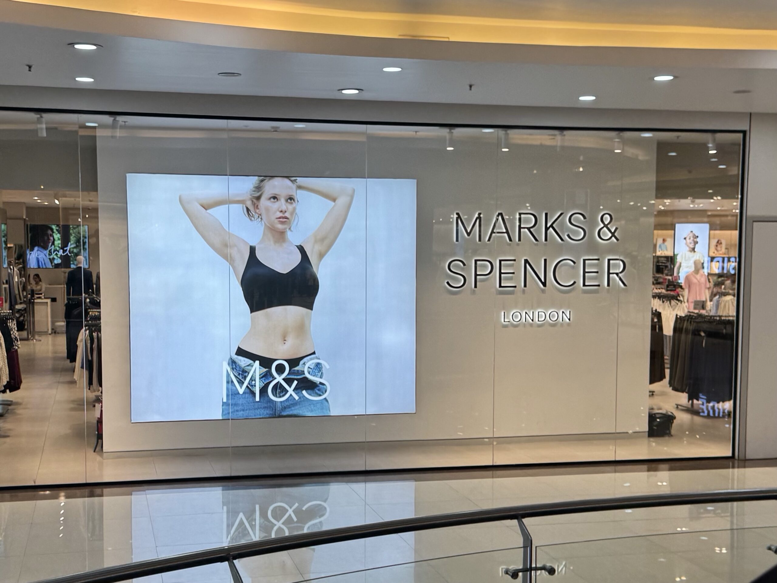 Front of a Marks & Spencer storefront with a large poster of a fit woman in a black sports bra and jeans, store interior visible behind glass doors.