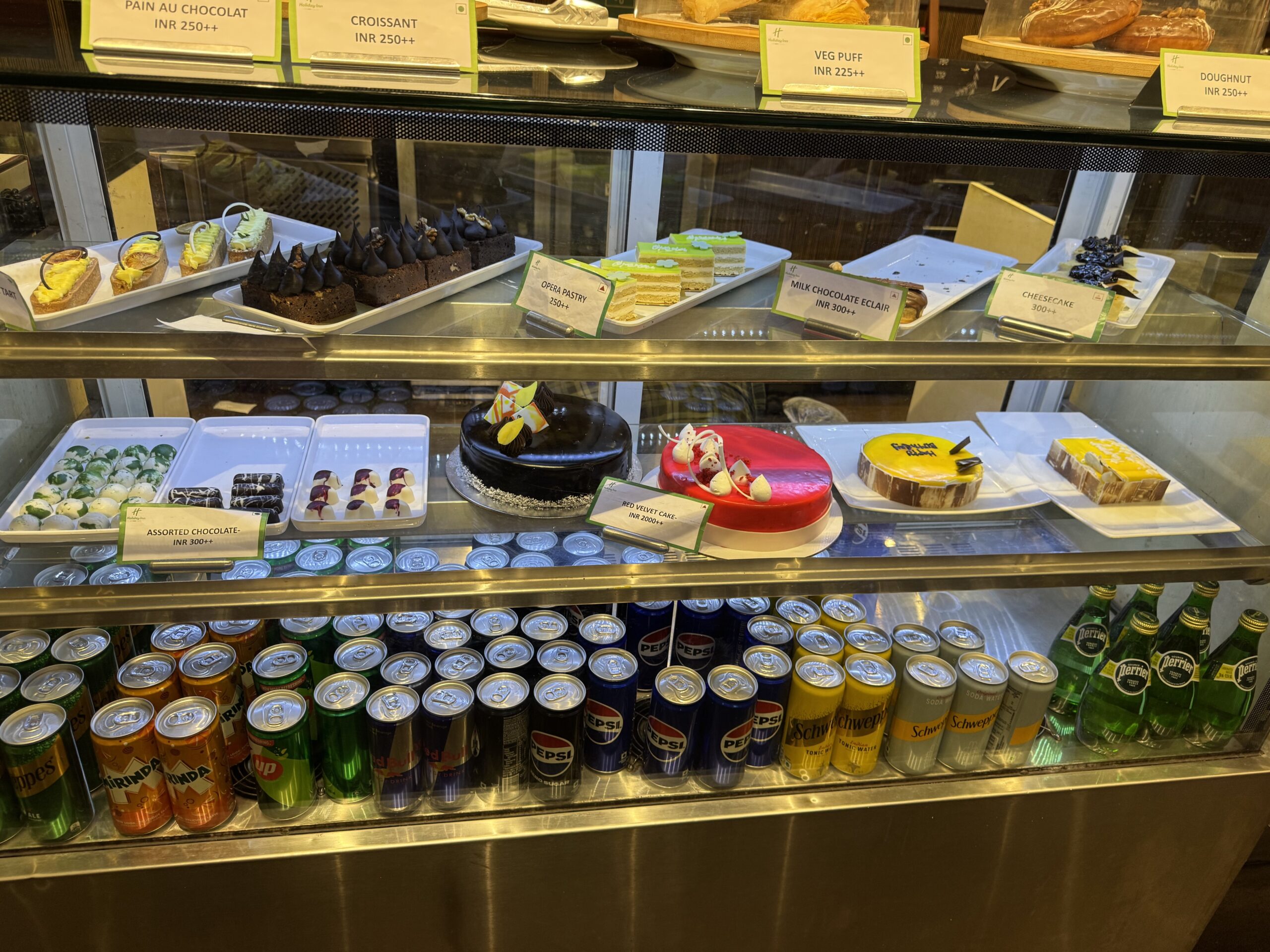 Display case with assorted pastries and cakes on white trays, labels visible above and a row of canned drinks below.