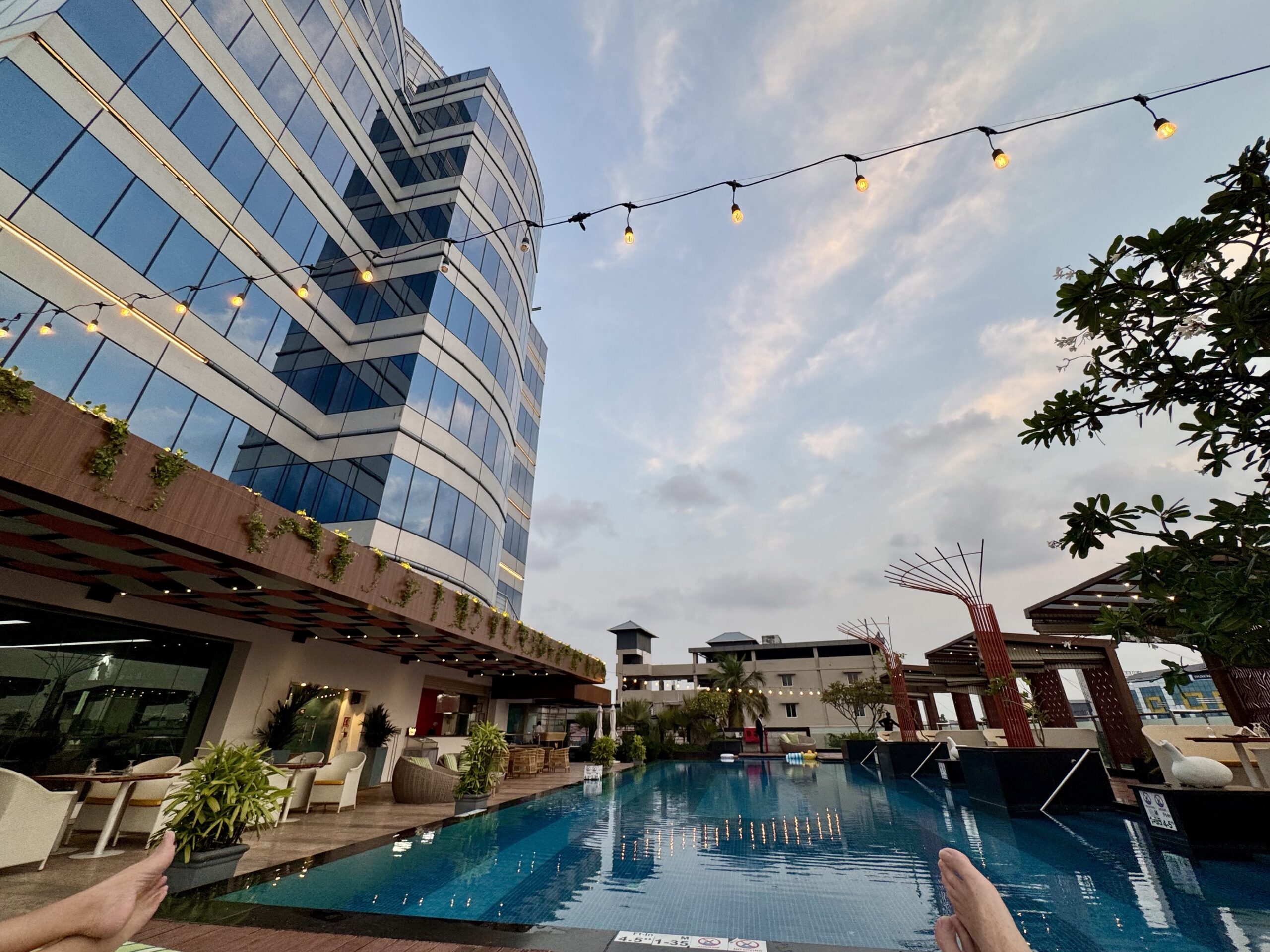 First-person poolside view with feet in the foreground, modern glass building on the left, string lights above, and a clear blue sky.