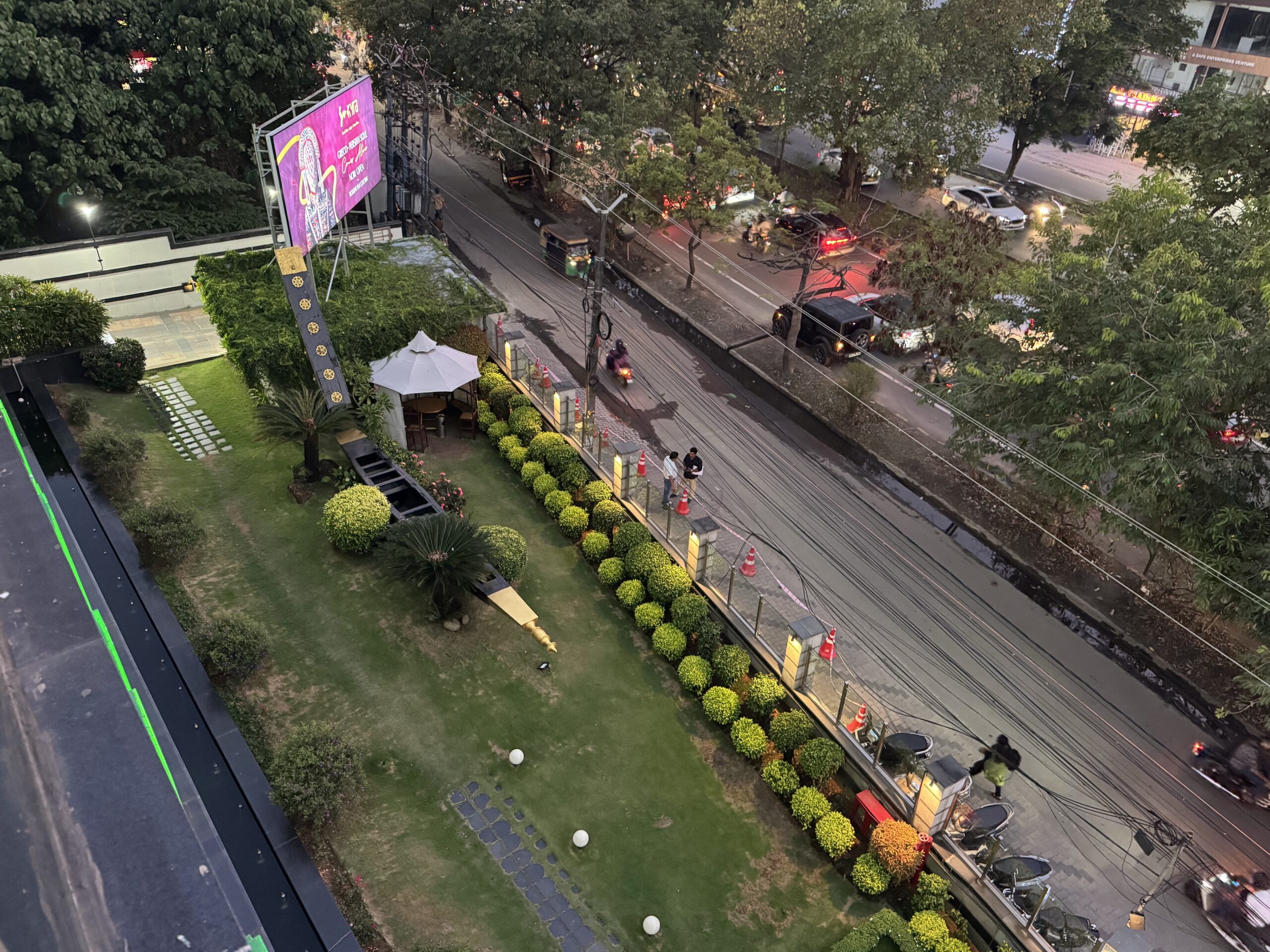 Aerial view of a street scene: cars, motorcycles, and a tuk-tuk pass by, with trees lining the road and a purple billboard on the left. A manicured lawn and cafe seating with a white umbrella are in the foreground.