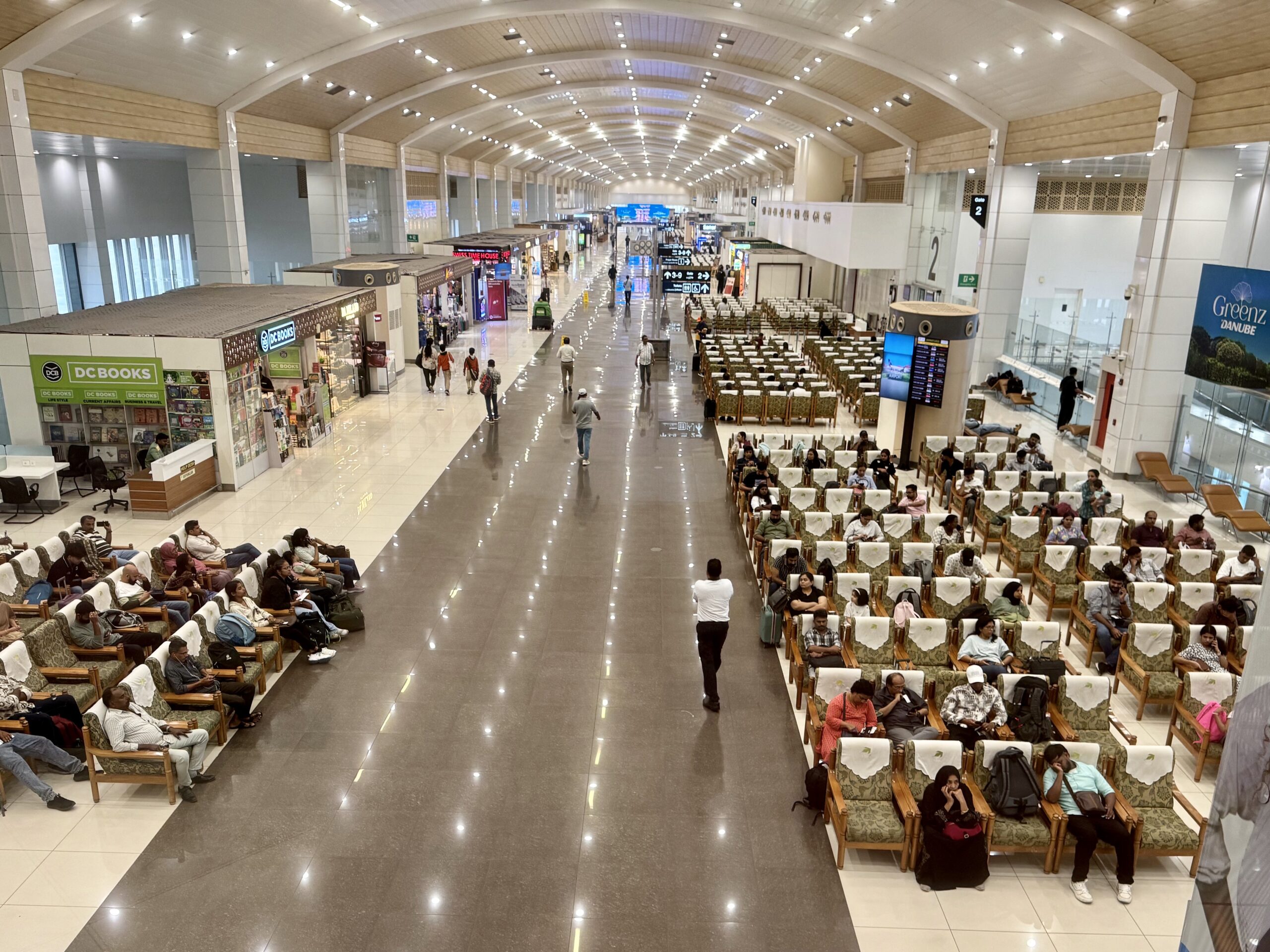 Wide interior of a spacious airport terminal with rows of seating and travelers waiting near shops on the left.