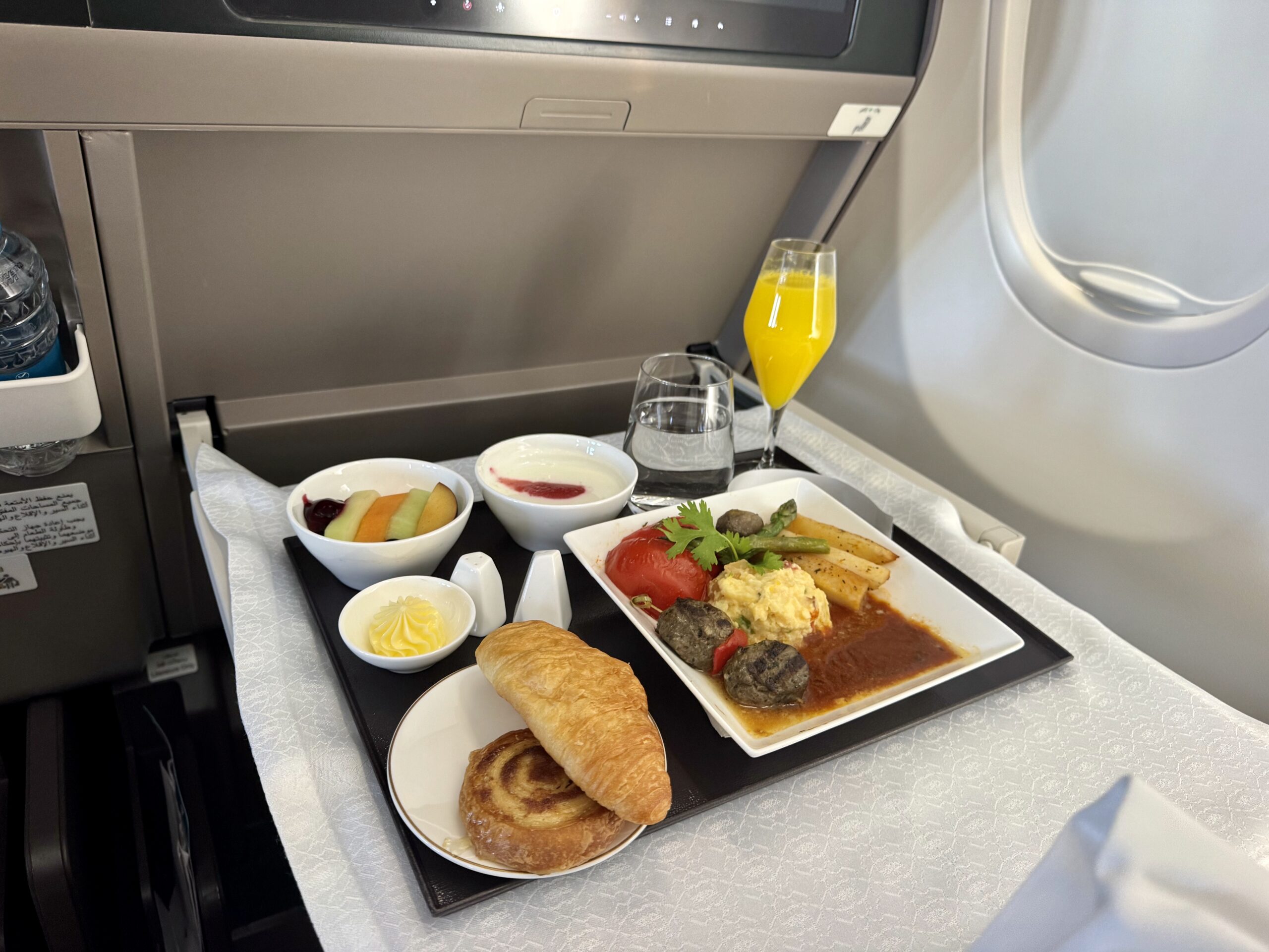 In-flight meal on a tray: croissant, bread roll, fruit bowl, yogurt with berry sauce, a main plate with vegetables and sauce, and a glass of orange juice on the tray table.