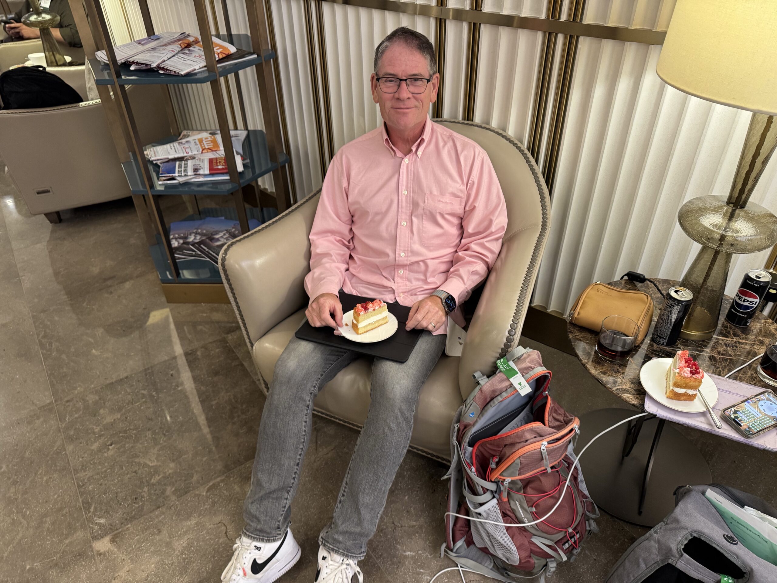 Man in a pink button-down shirt sits in a beige chair, holding a plate with a slice of cake.