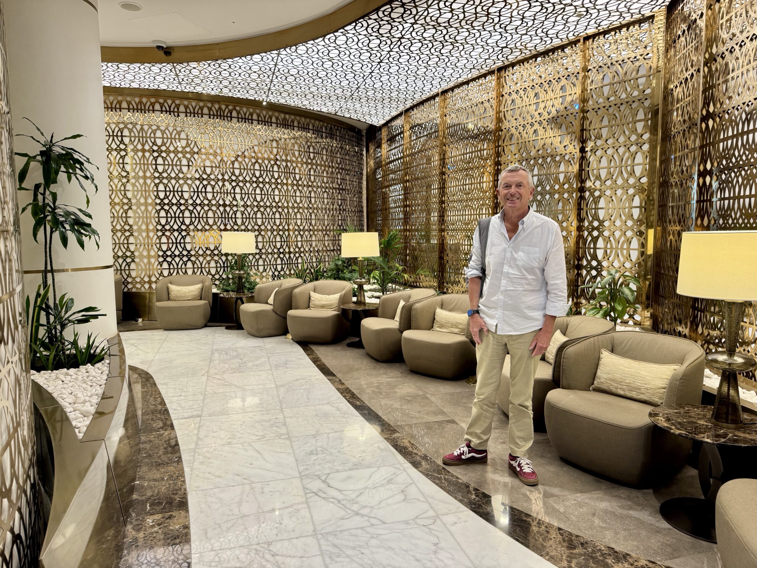 A man in a white shirt and khaki pants stands in a luxurious hotel lobby with gold lattice walls and tropical plants behind him.