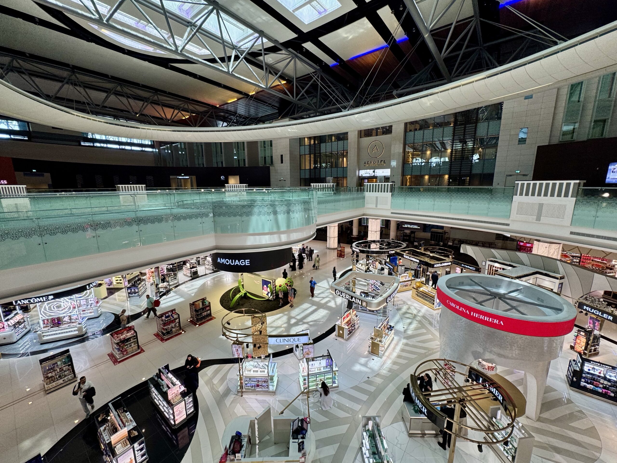 Interior of a modern multi-story shopping mall with glass railings, storefronts (Lancôme, Amouage, Clinique) and a central circular escalator/viewing area.