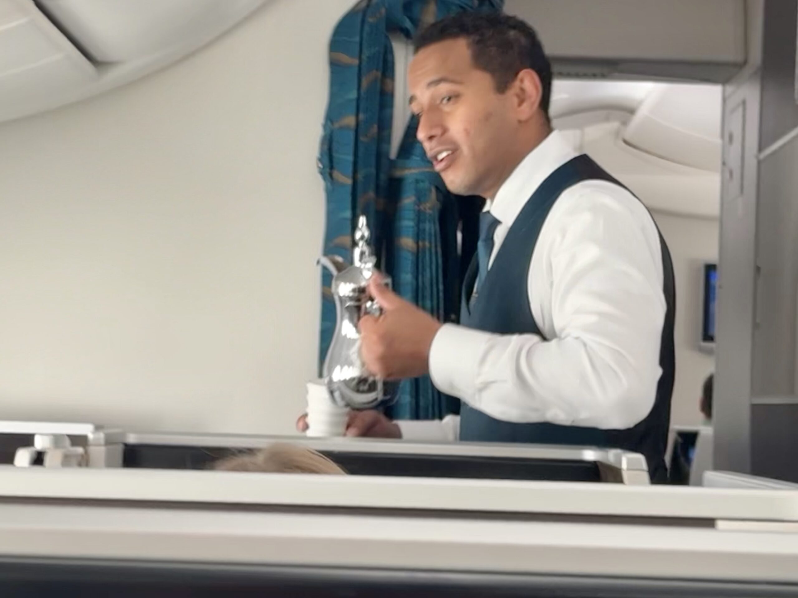Flight attendant in a teal vest serving a drink on an airplane, holding a metal pitcher and glass.