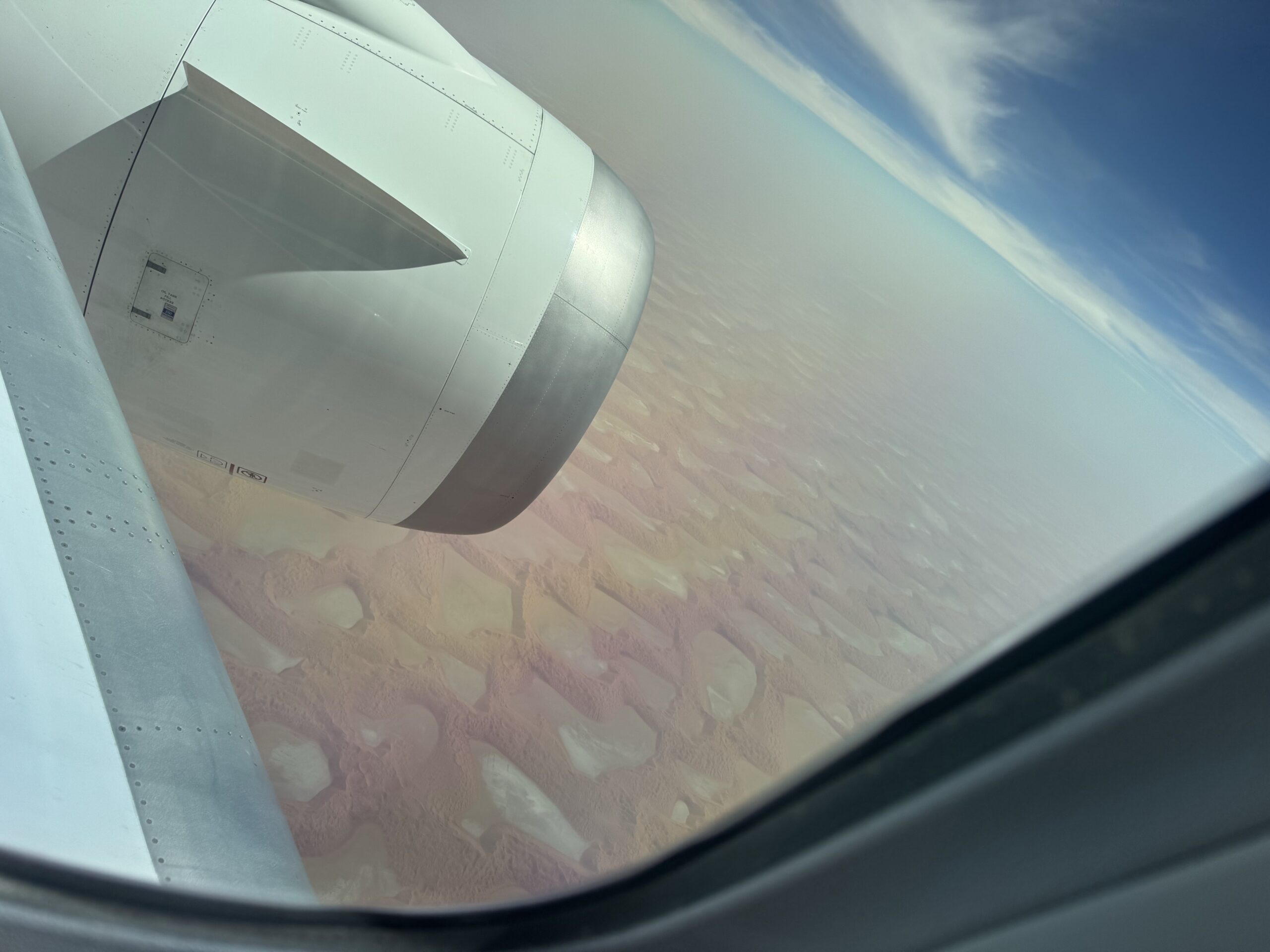 View from airplane window showing the wing and engine nacelle with a desert landscape below and a blue sky above
