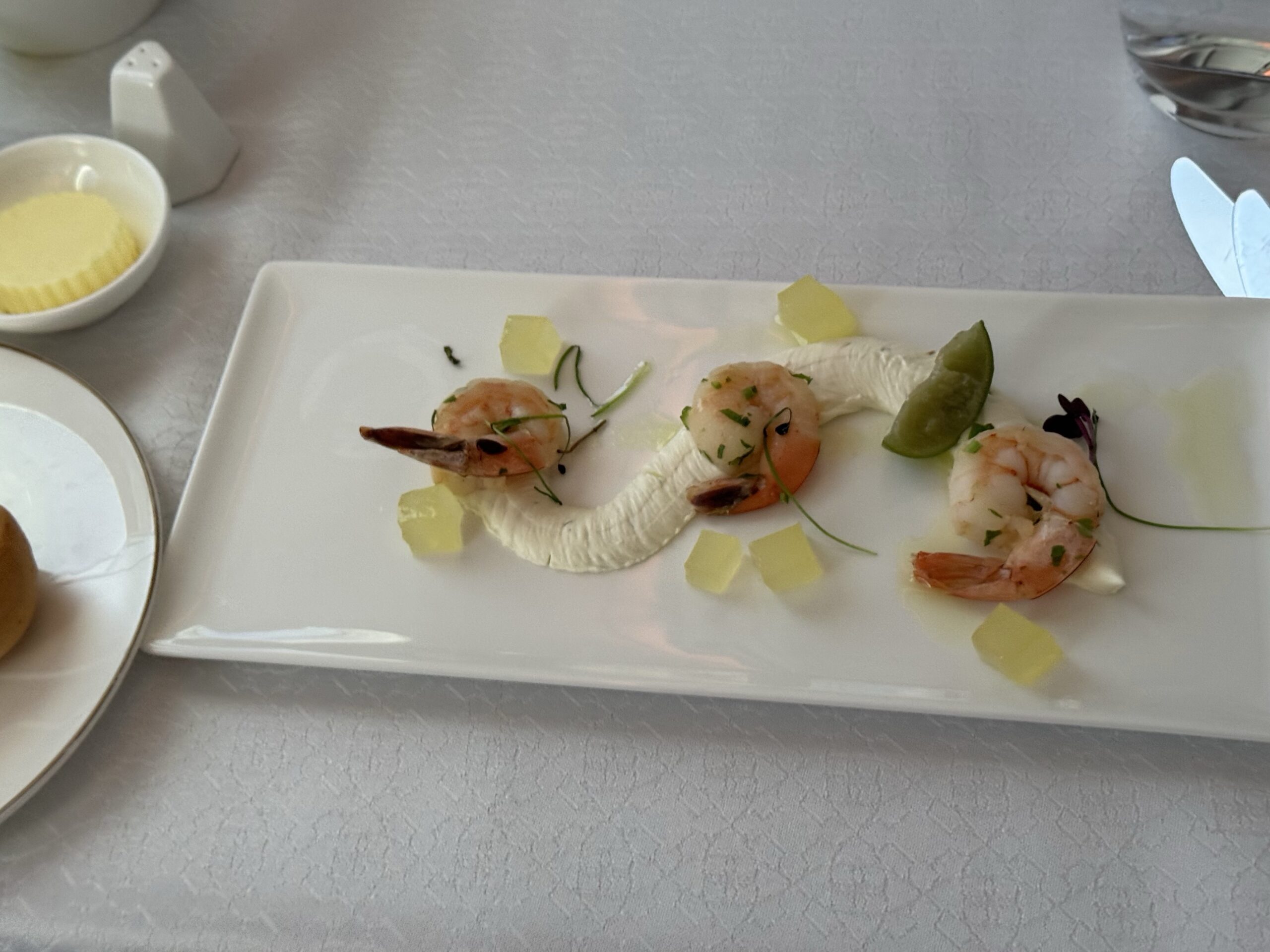 Shrimp arranged on a white rectangular plate with lime wedge, herb garnish, and small yellow jelly cubes; butter bowl visible nearby in a fine-dining setting.