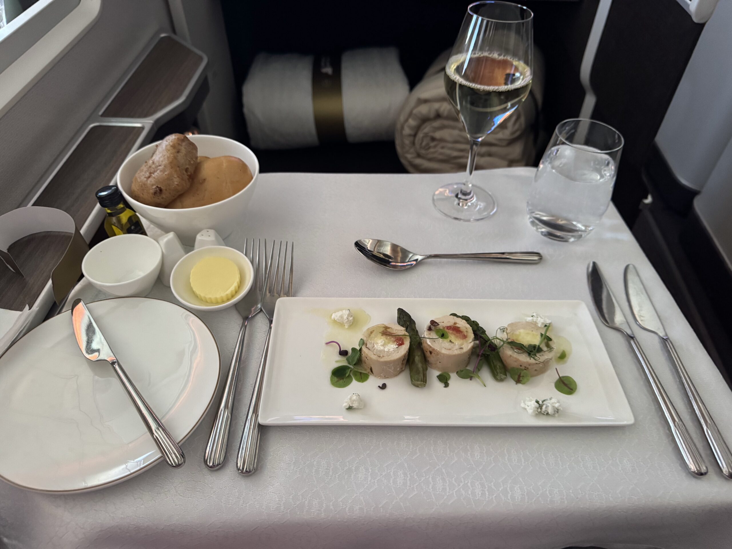 In‑flight meal on a tray: rectangular plate with roulade pieces, greens and drizzle; bread rolls in a bowl with butter, alongside a wine glass and a water glass, plus cutlery and condiments.