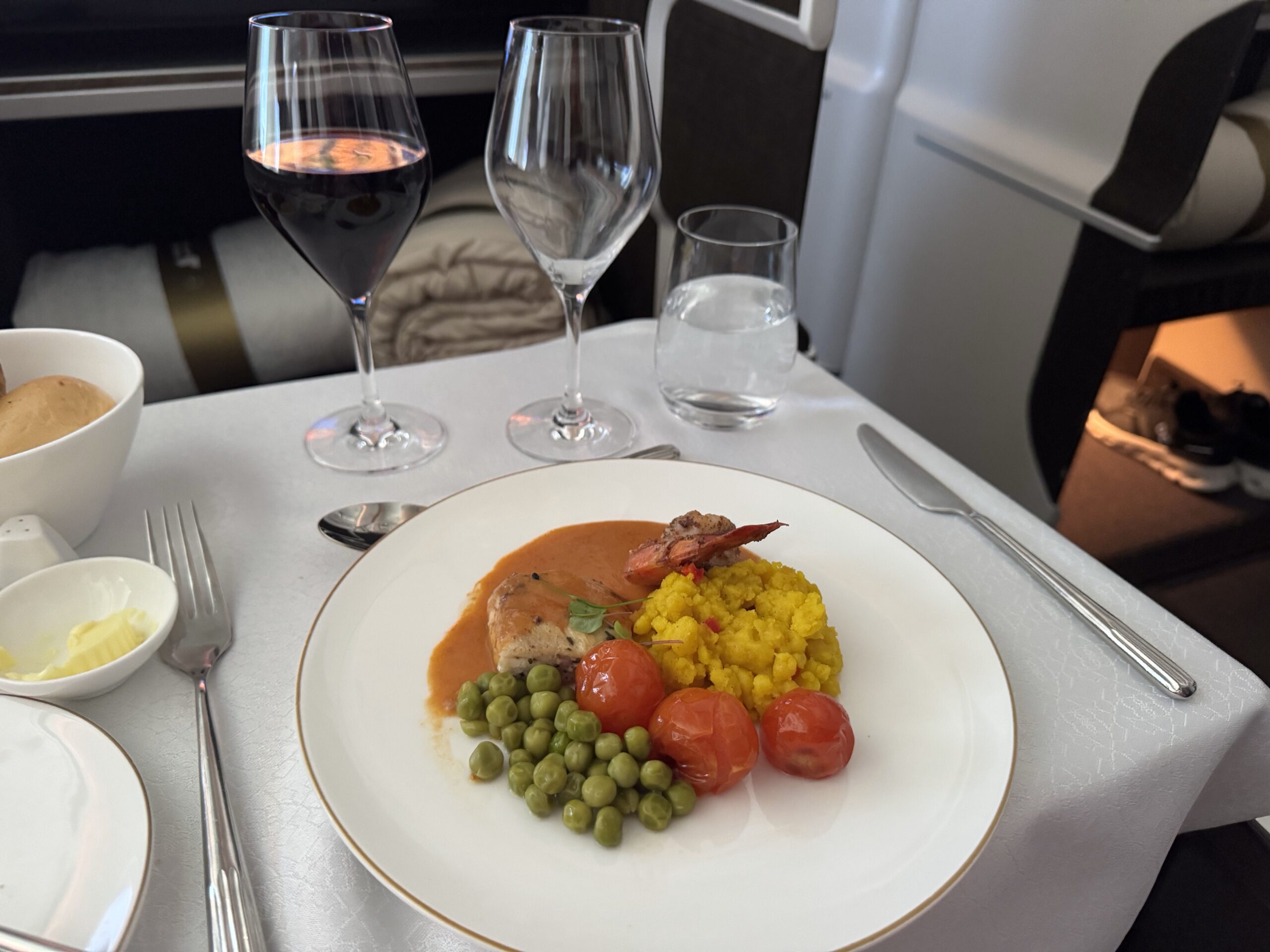 Plate of shrimp with yellow rice, cherry tomatoes, and green peas; two wine glasses and a water glass on a white table in an airplane cabin.