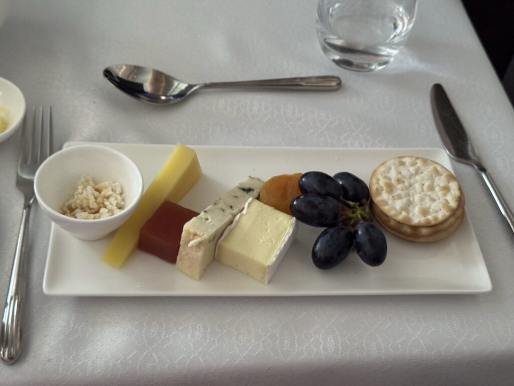 Cheese board with assorted cheeses (blue, Brie), yellow cheese, red fruit jelly slice, green grapes, crackers, and a small bowl of crumbled topping on a white plate.