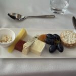 Cheese board with assorted cheeses (blue, Brie), yellow cheese, red fruit jelly slice, green grapes, crackers, and a small bowl of crumbled topping on a white plate.