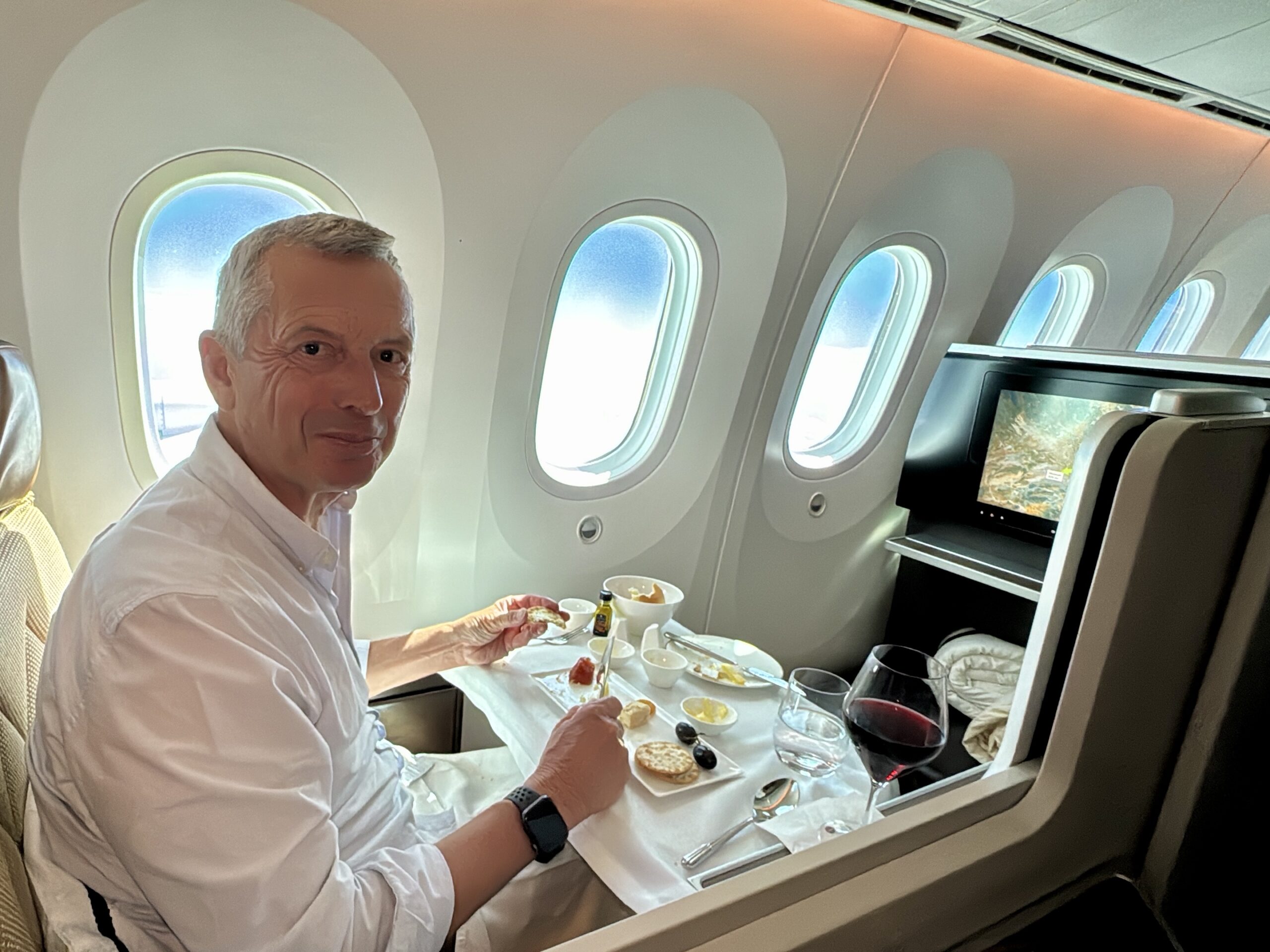 Man dining in a first-class airplane cabin, with a food tray, glass of red wine, and window seats in the background.