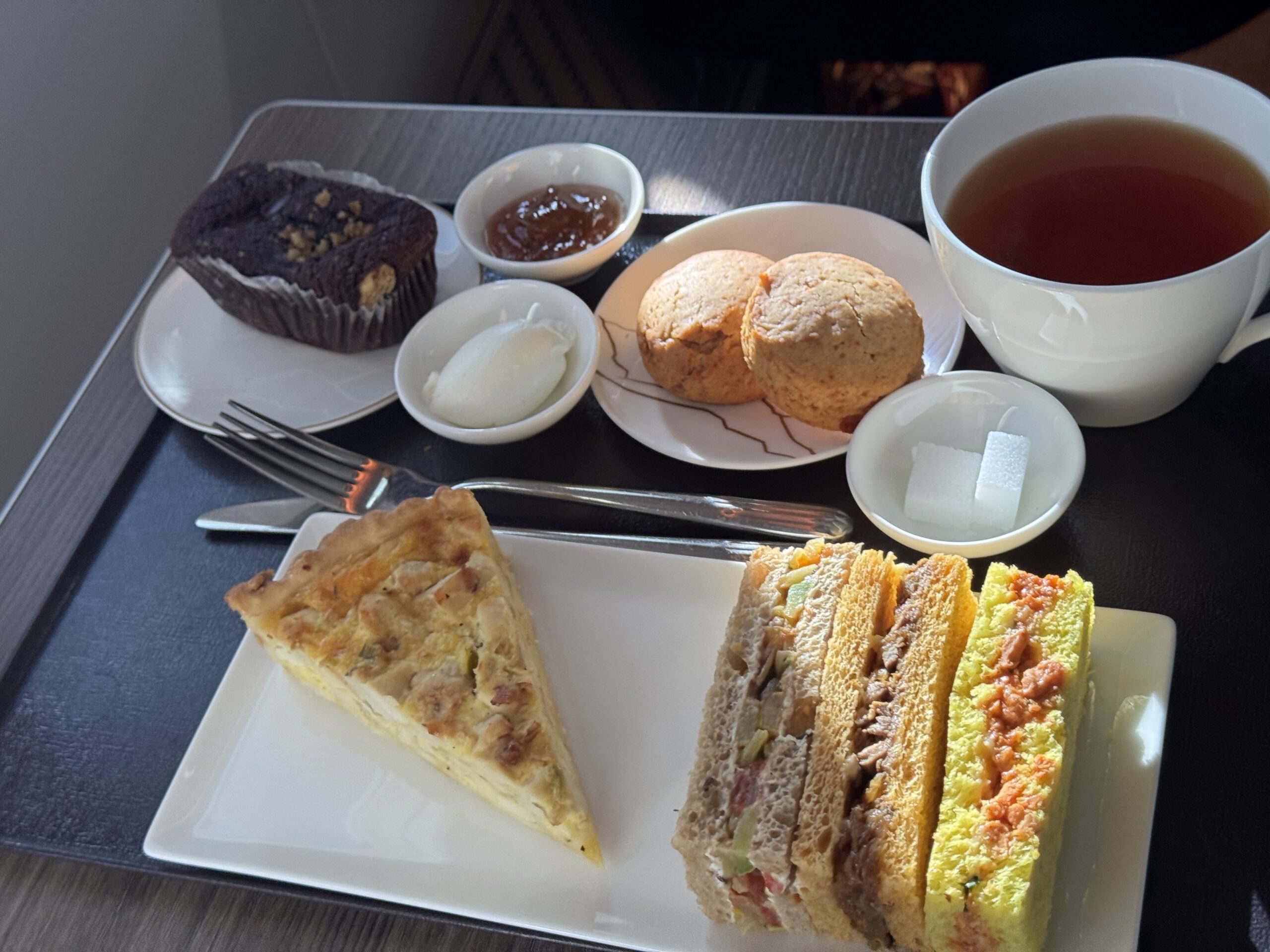Assortment of tea-time treats on a tray: chocolate muffin with nuts, jam, vanilla ice cream, two scones, and colorful finger sandwiches with a cup of tea nearby.