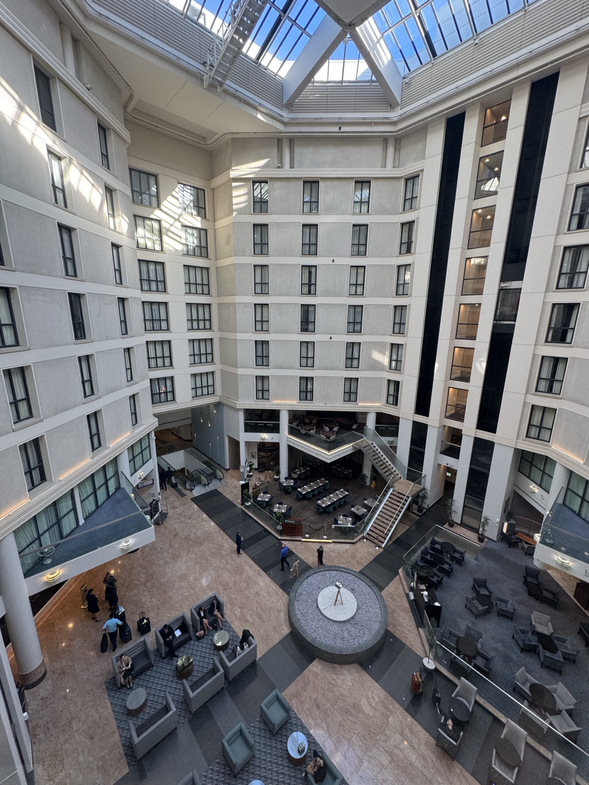 Hotel atrium with glass skylight, curved white walls, and multiple floors of windows surrounding a central lounge area.